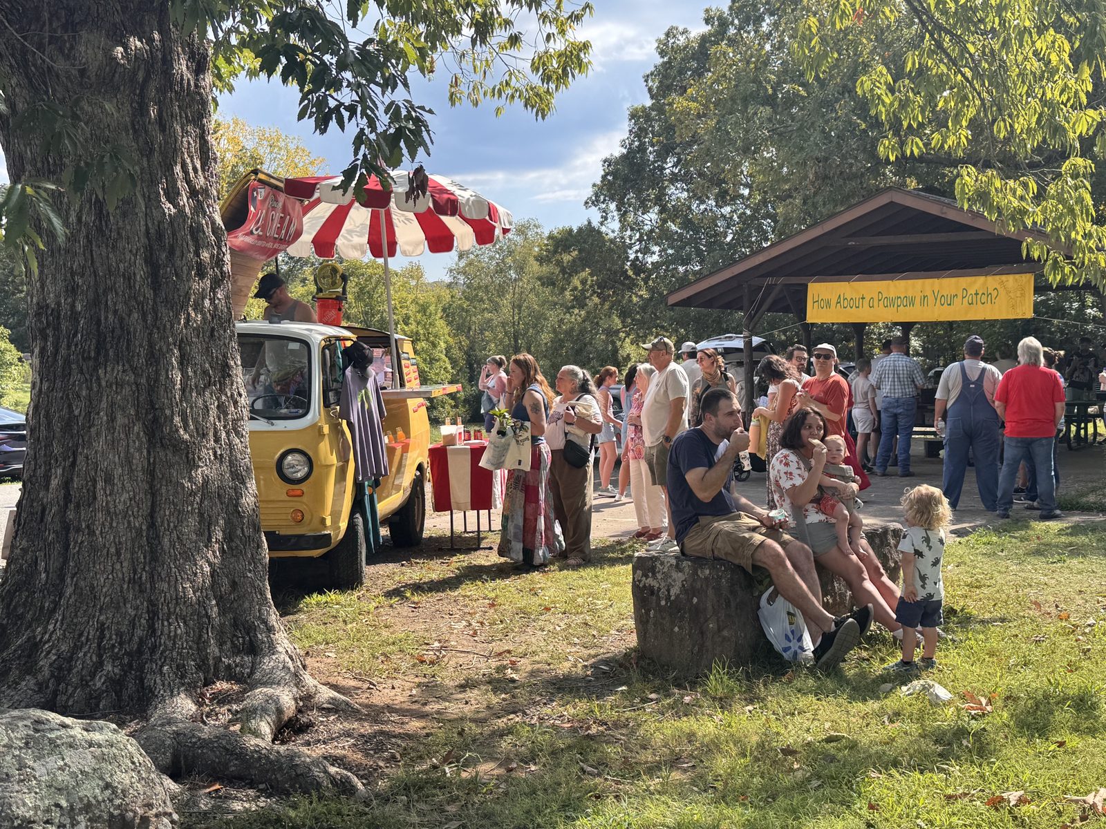 Sunlit crowd around a vintage yellow ice cream truck, a family with kids eating ice cream on a rock, festival banner behind